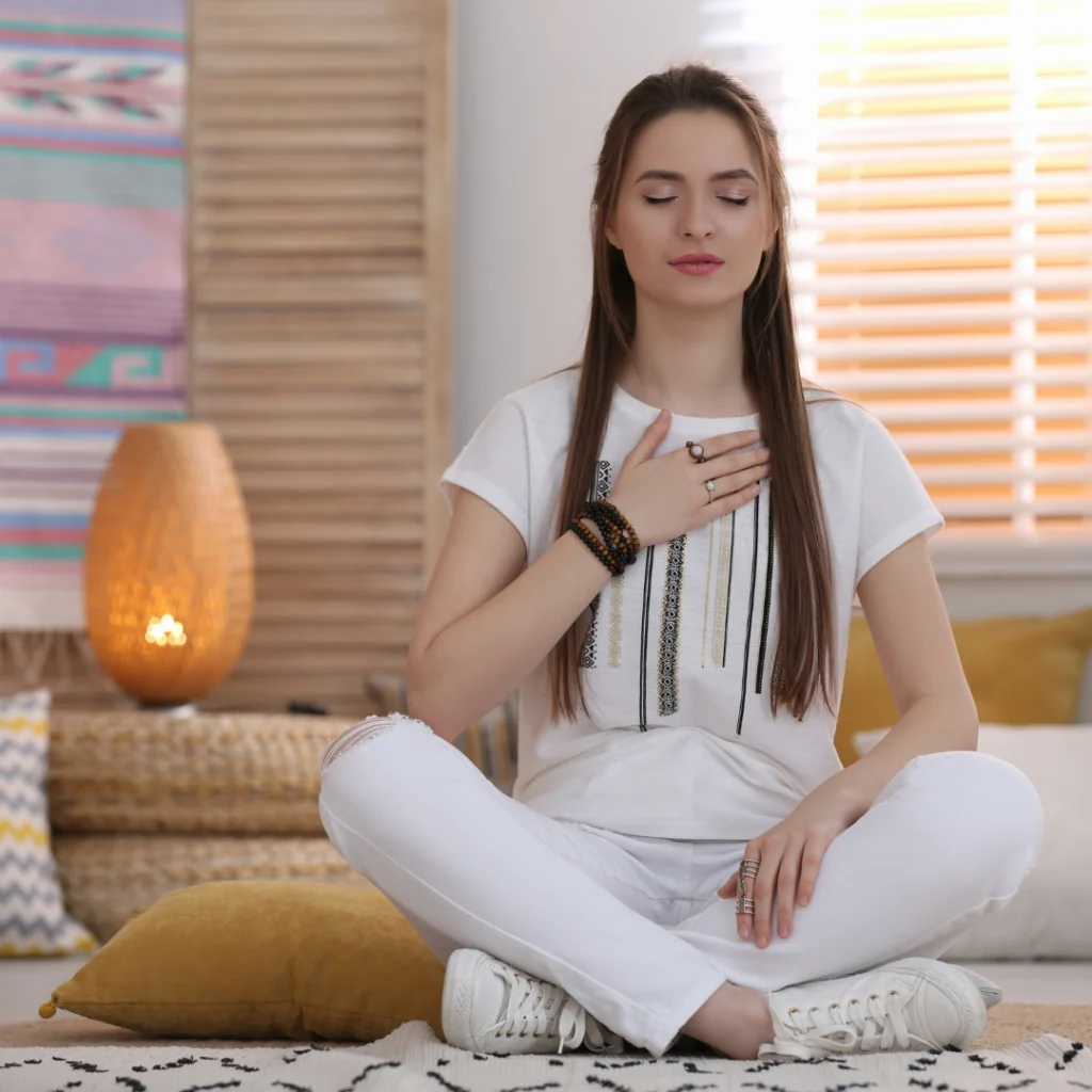 A woman with brown hair, dressed in all white, sits cross-legged on the floor with her hand over her heart.